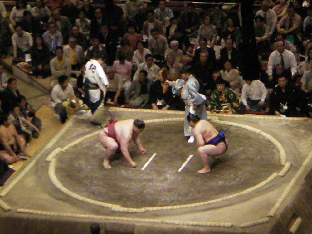 two wrestlers squatting and facing each other inside a white circle while the referee observes